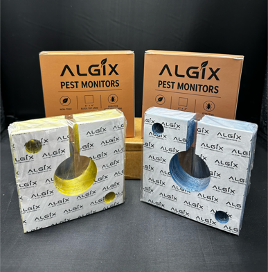 Two ALGIX pest monitor devices with their retail packaging boxes, shown on a dark studio background. The left device is yellow with a white patterned box and a gold circular window. Sticky Pest Trap Monitors 