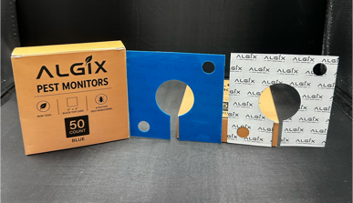 Two ALGIX pest monitor devices with their retail packaging boxes, shown on a dark studio background. The left device is blue with a white patterned box and a gold circular window. Sticky Pest Trap Monitors 