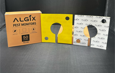 Two ALGIX pest monitor devices with their retail packaging boxes, shown on a dark studio background. The left device is yellow with a white patterned box and a gold circular window. Sticky Pest Trap Monitors 